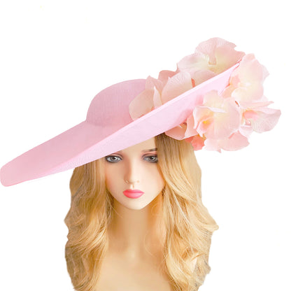 A light pink wide-brimmed extra large fascinator hat with large layered fabric flowers suitable for a wedding guest